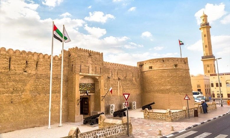 Dubai Museum exterior with traditional fort walls, cannons, UAE flags, and a mosque in the background.