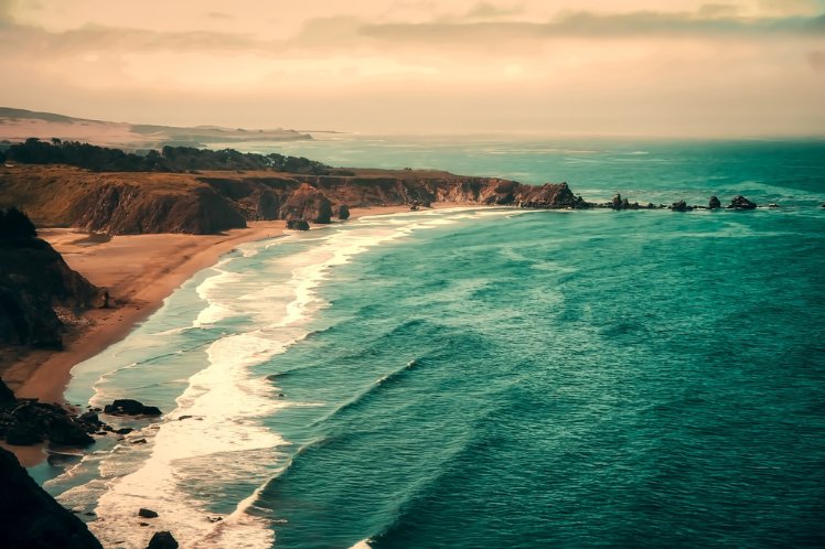 California Coastline with cliffs, beach, and waves, under a hazy sky.