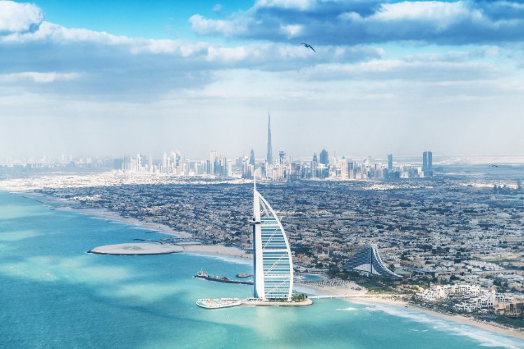 Aerial view of Dubai coastline with Burj Al Arab in the foreground and city skyline in the background.